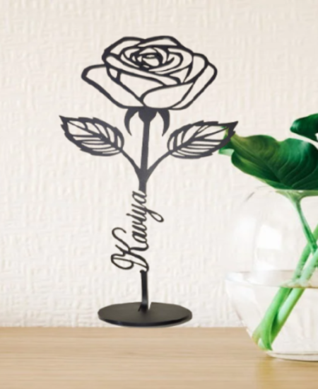 Black metal rose sculpture with 'Kavya' engraved on a wooden surface with a white wall background.